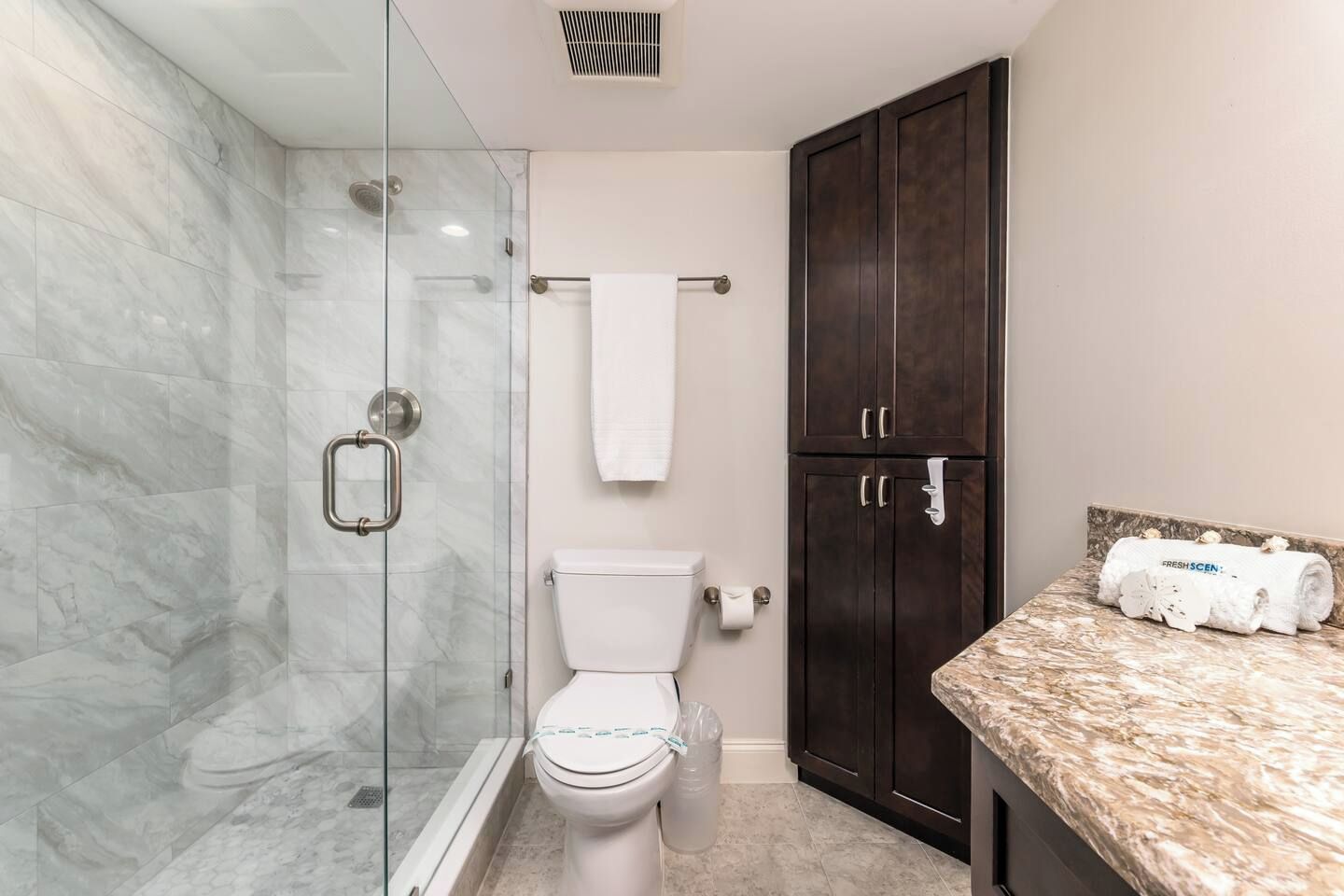 A bathroom with a glass-enclosed shower, a toilet, a dark wooden cabinet, and a marble countertop.