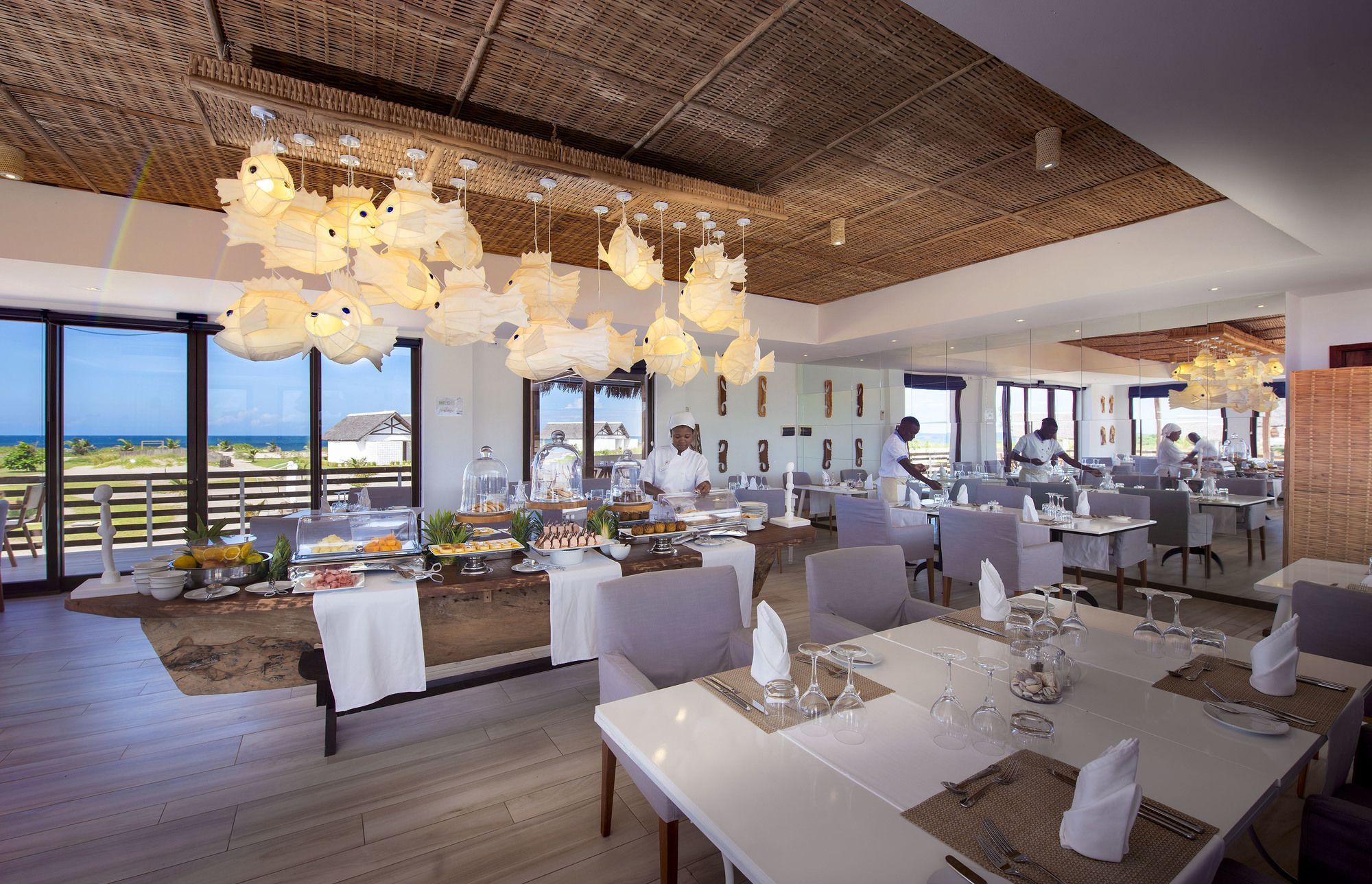 A restaurant with a view of the ocean, featuring a high ceiling with hanging lights, tables set for dining, and a chef preparing food.