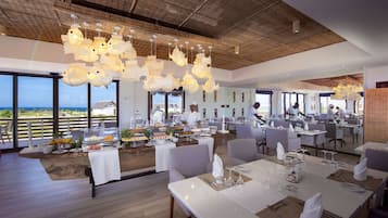 A restaurant with a view of the ocean, featuring a high ceiling with hanging lights, tables set for dining, and a chef preparing food.