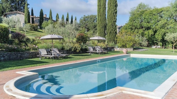 A rectangular swimming pool with a curved edge, surrounded by a brick patio. There are lounge chairs and umbrellas in the background. - La Pieve Marsina (Gaiole in Chianti)