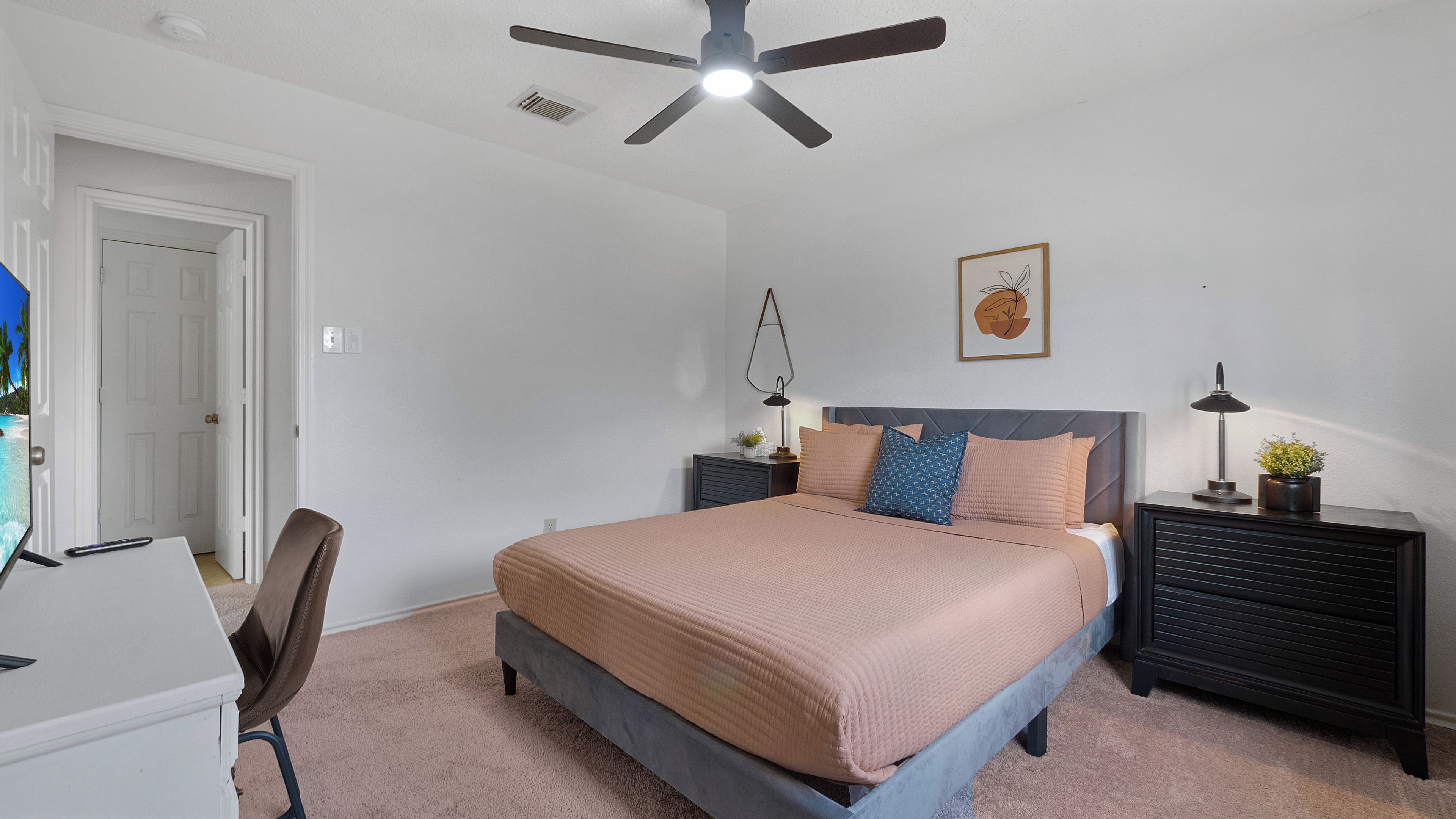 Ein Schlafzimmer mit Bett, Nachttischen, Schreibtisch, Stuhl, Deckenventilator und einer Tür.