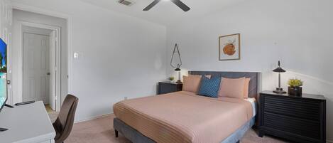 Un dormitorio con cama, mesitas de noche, un escritorio, una silla, un ventilador de techo y una puerta.