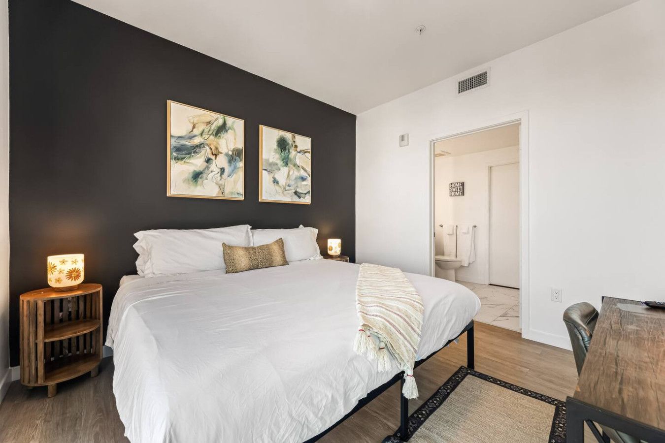 Quarto moderno com uma cama grande, duas pinturas abstratas penduradas na parede, uma luminária de cabeceira e uma mesinha de madeira.