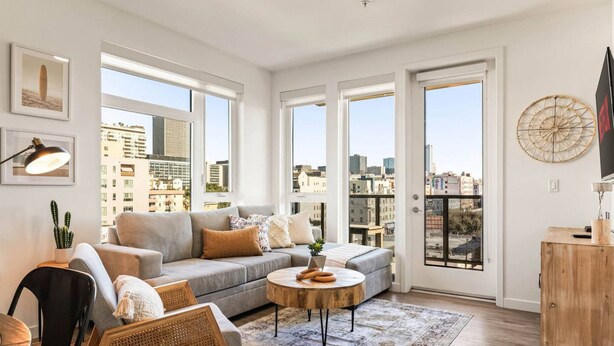 Una sala de estar moderna con un sofá gris, una mesa de centro redonda y vistas de la ciudad a través de grandes ventanales.