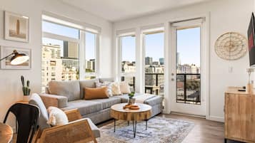 Un salon moderne avec un canapé gris, une table basse ronde et une vue sur la ville grâce à de grandes fenêtres.