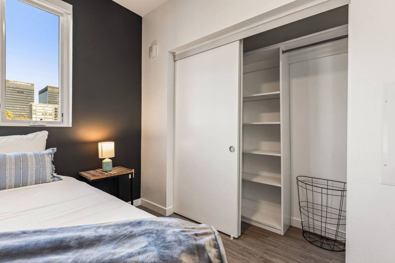Une chambre à coucher avec un lit, une table de chevet avec une lampe, une armoire et une vue sur les bâtiments par la fenêtre.