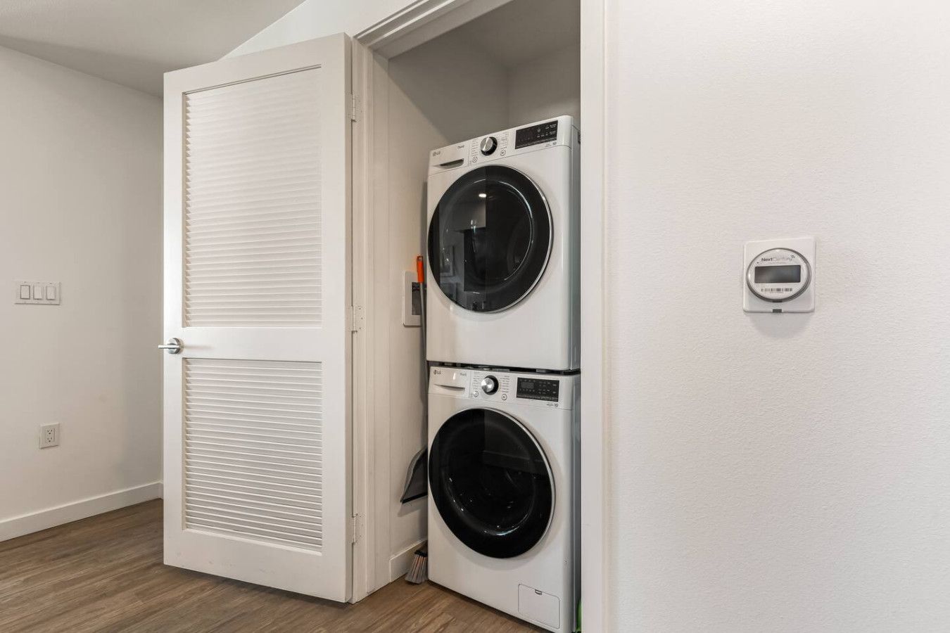 Une buanderie équipée de deux machines à laver à chargement frontal superposées, d’une porte blanche à persiennes et d’un minuteur fixé au mur.