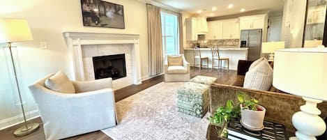 A living room with a fireplace, a ceiling fan, and a kitchen in the background.