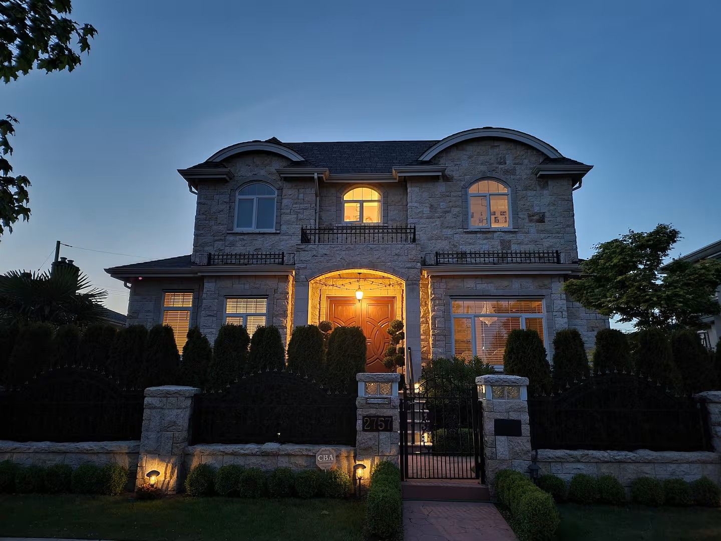 A two-story stone house with a central entrance, a gated driveway, and a well-maintained front yard with trimmed hedges.