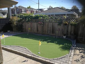 A backyard with a putting green, a wooden fence, and a house in the background. - Two bedrooms basement suite (Vancouver)