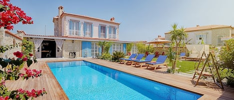 A swimming pool with lounge chairs, a stone building with blue shutters, and a clear sky.