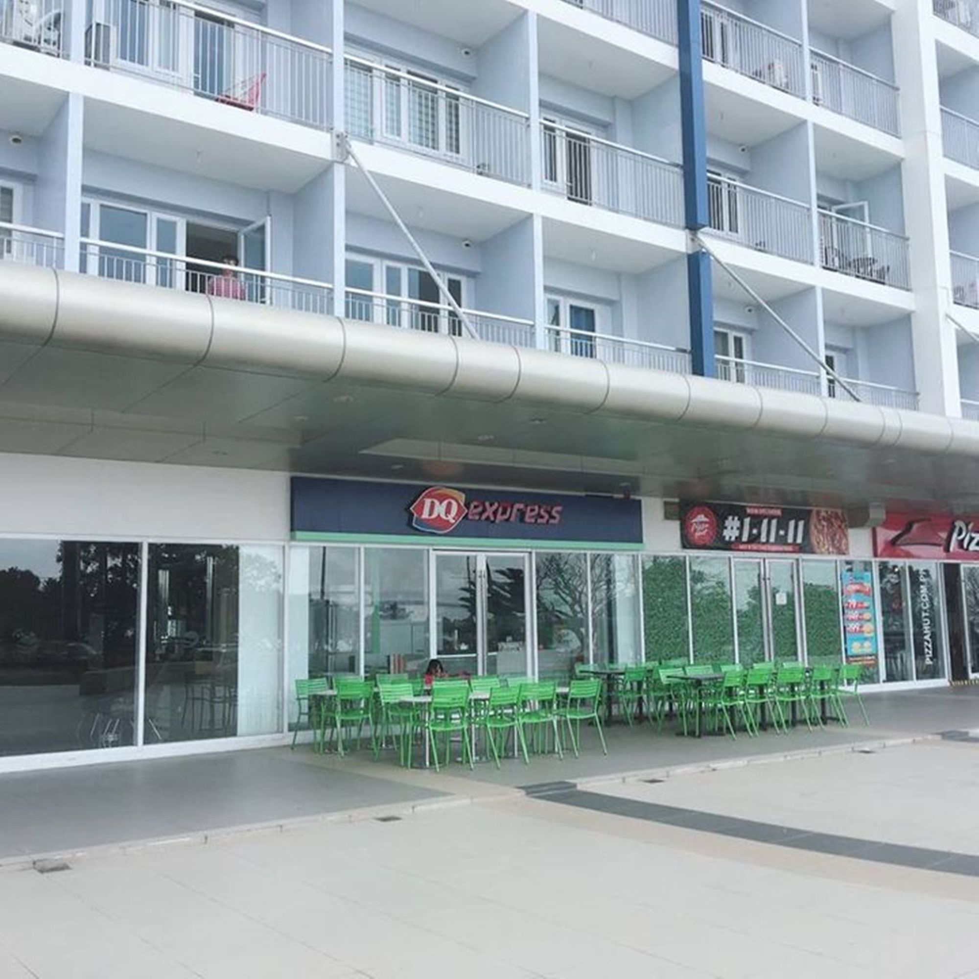 A multi-story building with a DQ Express restaurant on the ground floor, green chairs outside, and a Pizza Hut across the street.