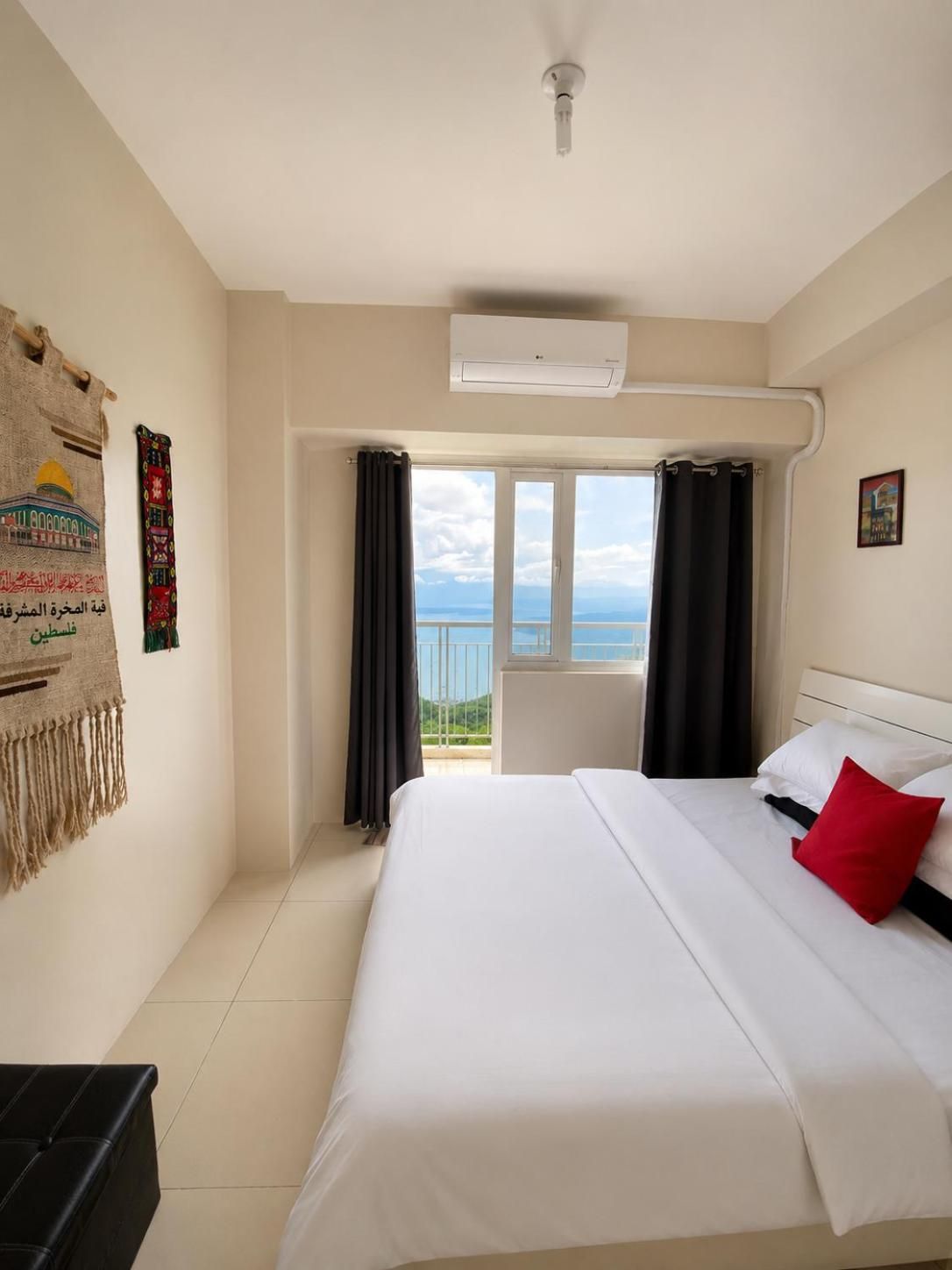 A hotel room with a bed, a ceiling fan, an air conditioning unit, a balcony with curtains, and a wall with a decorative tapestry.