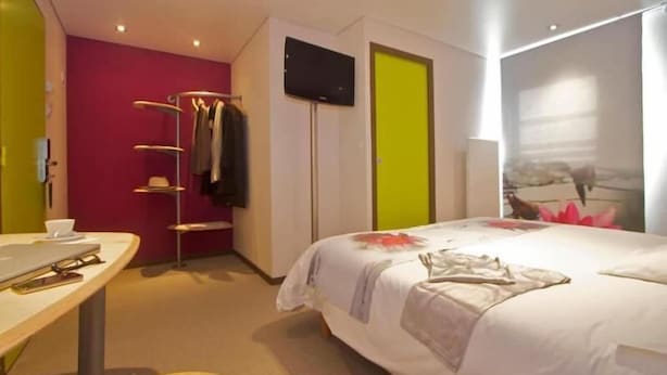 Quarto de hotel com cama, televisão, cabideiro e escrivaninha.