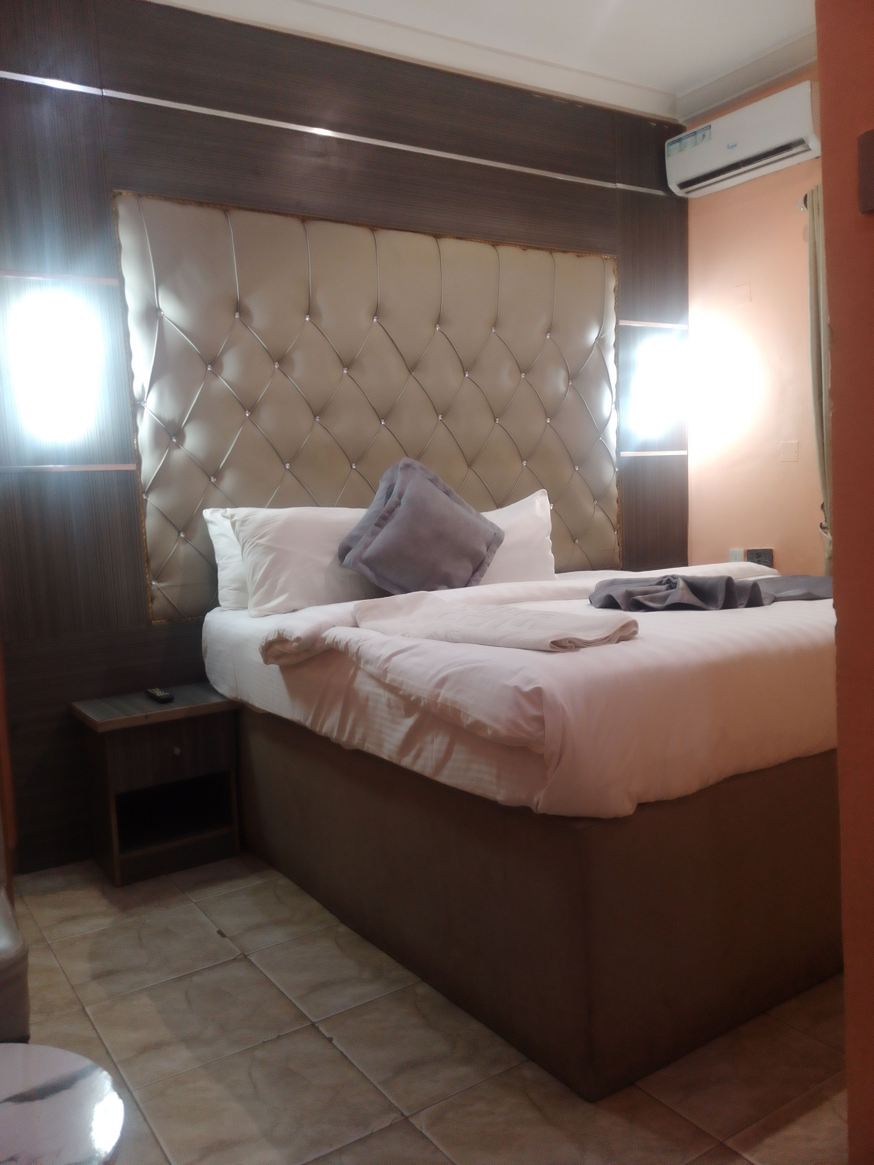 A bedroom with a tufted headboard, a bed with white bedding, a bedside table, and an air conditioning unit.