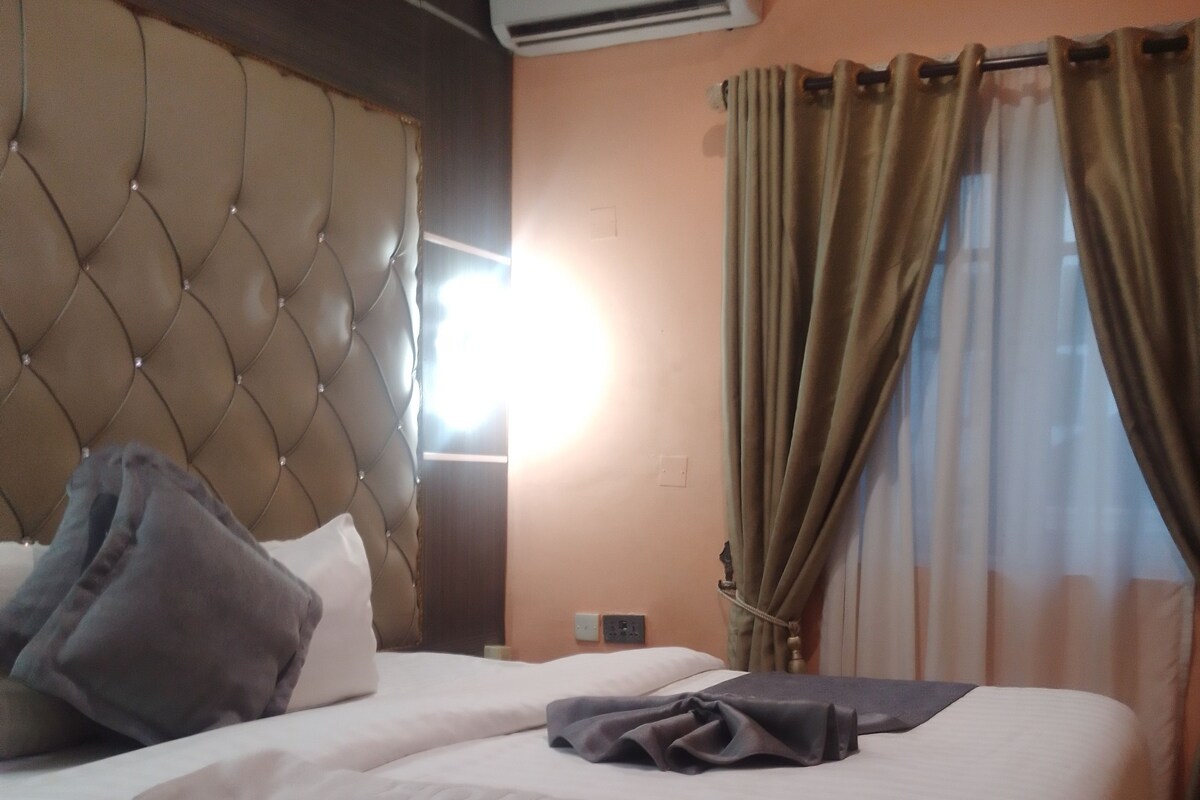 Een hotelkamer met een gestoffeerd hoofdeinde, een airconditioner, een bed met witte lakens, een nachtkastje en gordijnen.