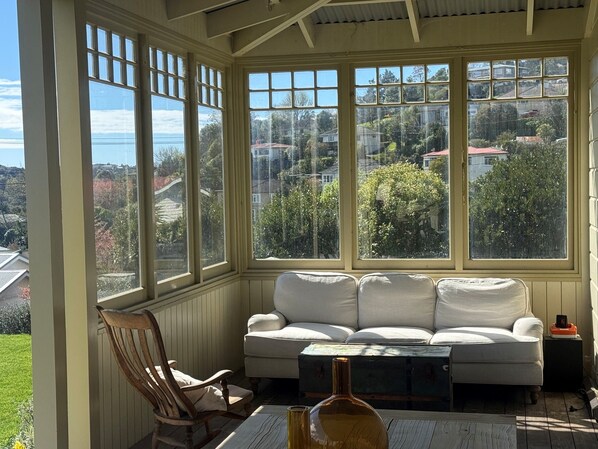 A conservatory with a white sofa and a wooden chair, a table with a bottle, and a view of a residential area.