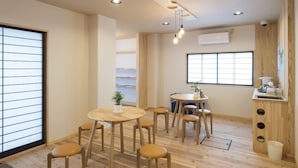 Une pièce avec un plancher en bois, une table ronde entourée de chaises, une fenêtre avec des stores, un plafond doté d’un éclairage encastré et une petite plante posée sur la table.