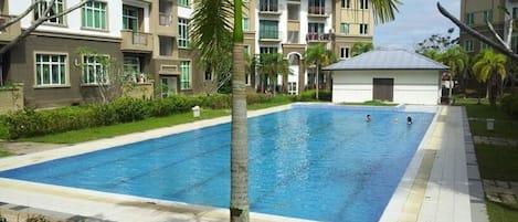 A rectangular swimming pool surrounded by a paved walkway, with a multi-story building in the background.