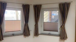 Una habitación con tres ventanas, cada una con cortinas recogidas, mostrando una vista de un edificio.