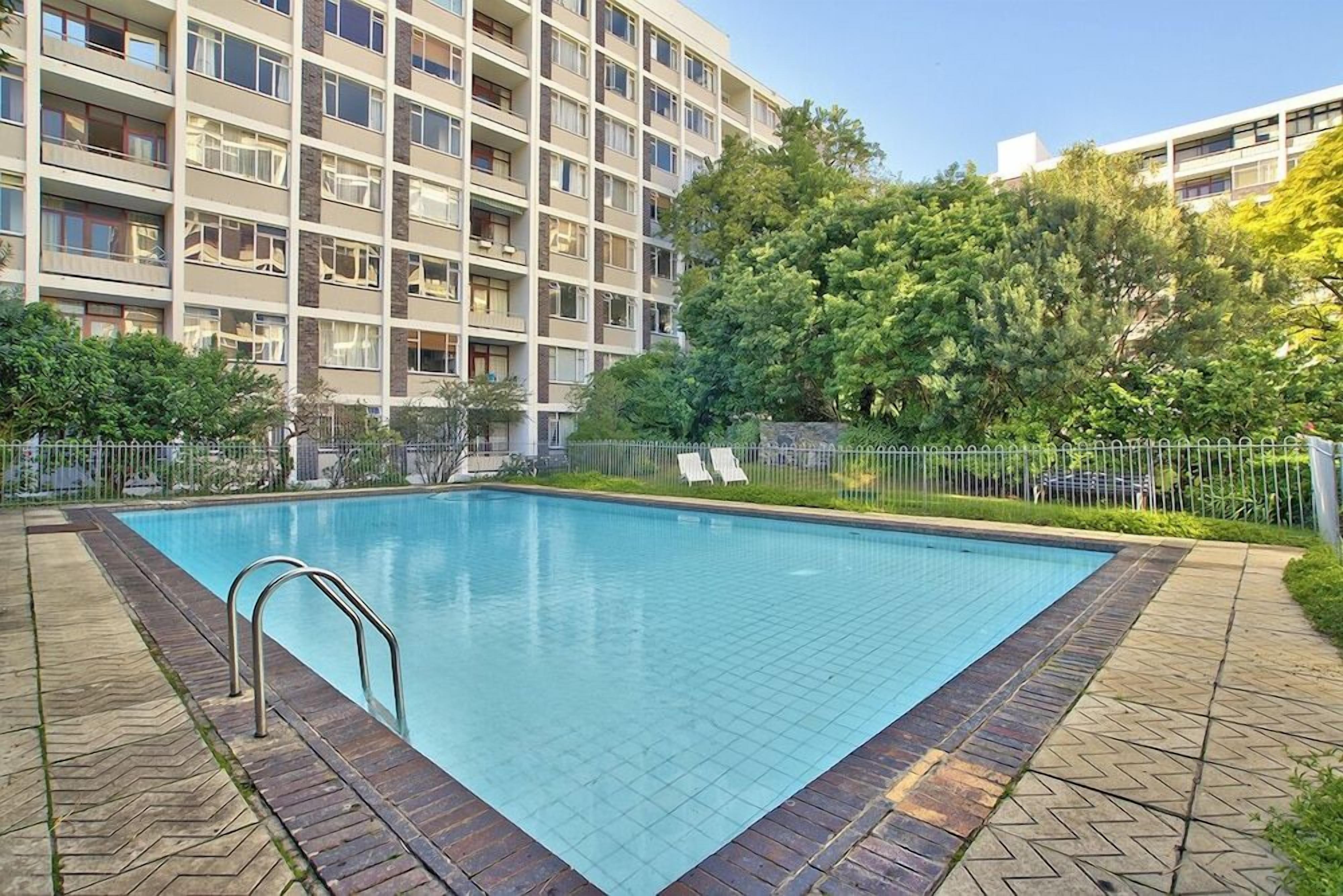 A swimming pool with a tiled deck, surrounded by a fence and trees, with a multi-story building in the background.