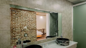 Une salle de bain avec un mur en briques, un miroir, deux vasques avec des cuves noires et un espace douche.