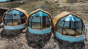 Three geodesic dome structures with transparent walls and thatched roofs, situated on rocky terrain. - Pacchanta Sky Dome Accommodation (Ocongate)