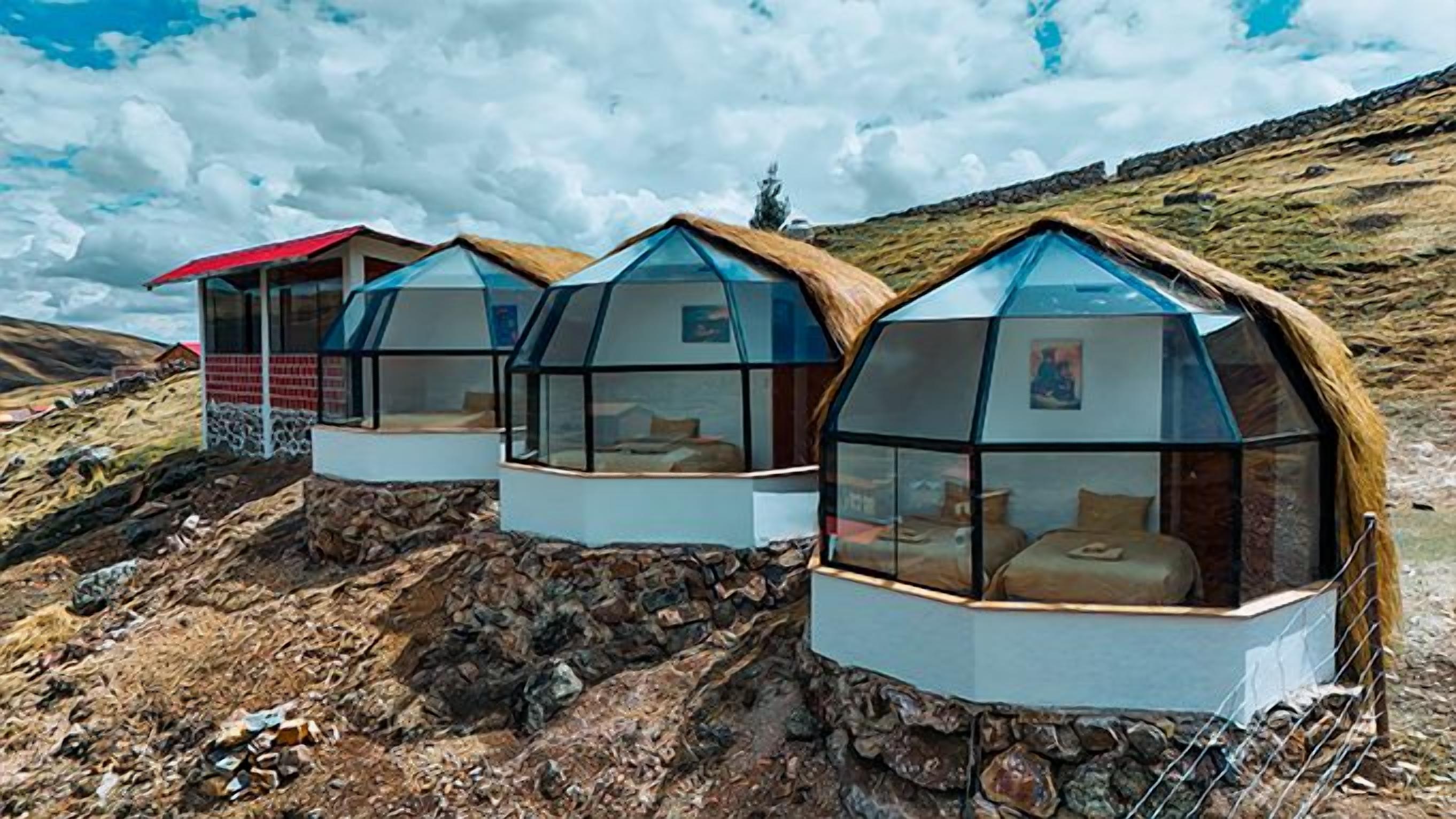 Three geodesic dome structures with glass walls and thatched roofs, situated on a rocky hillside.