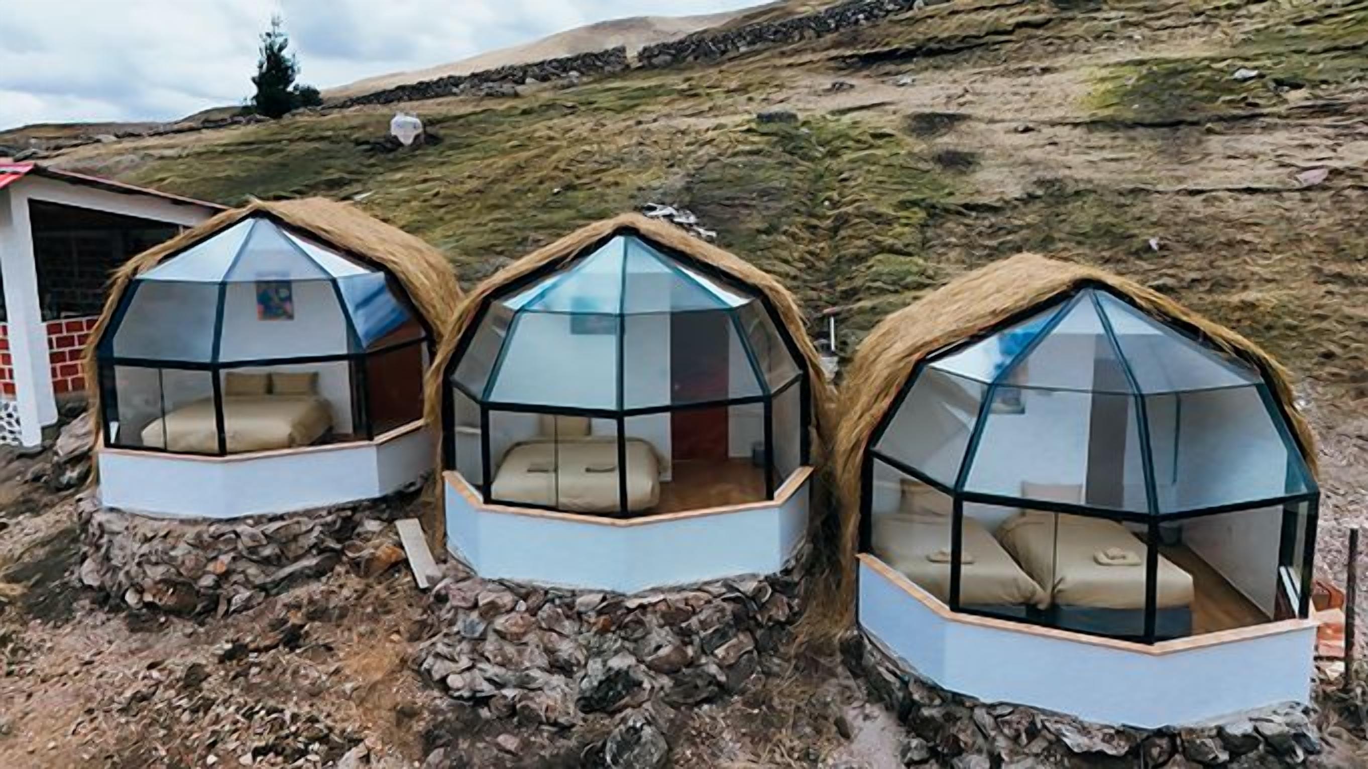 Tres estructuras de domos geodésicos con paredes traslúcidas y techos de paja, ubicadas en terreno rocoso con una colina cubierta de hierba al fondo.