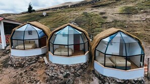 Tres estructuras de domos geodésicos con paredes traslúcidas y techos de paja, ubicadas en terreno rocoso con una colina cubierta de hierba al fondo.