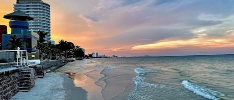 A beach with a curved hotel structure, a stone wall, and a sunset.
