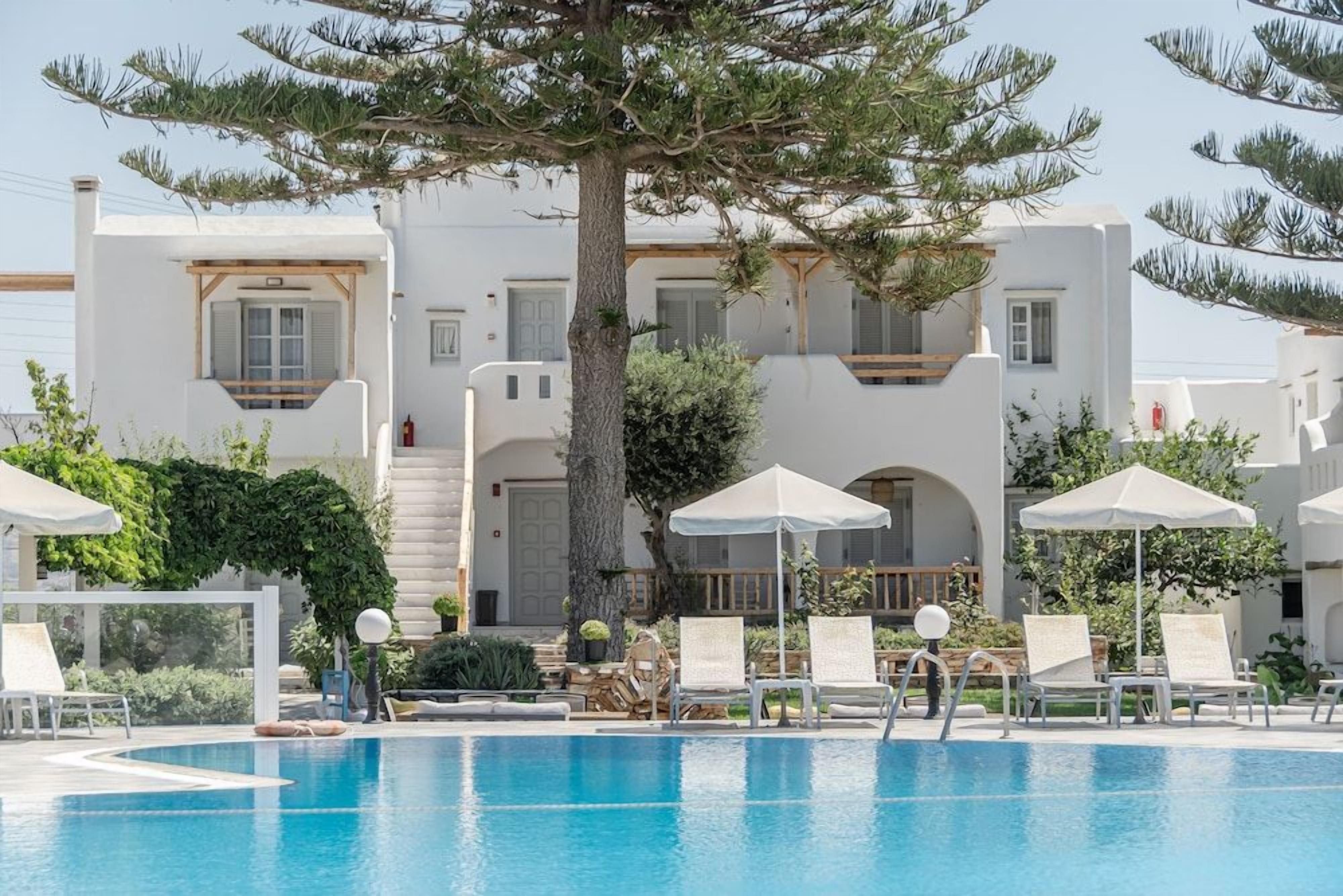 A pool area with lounge chairs and umbrellas, surrounded by white buildings with arched windows and a large tree.