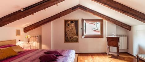 Un dormitorio con techo inclinado, vigas de madera, una cama con colcha estampada, una mesita de noche pequeña, un radiador y una ventana con vistas.