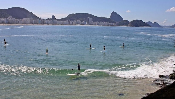 Paisaje costero con gente haciendo paddleboard, agua cristalina y un horizonte urbano de fondo.