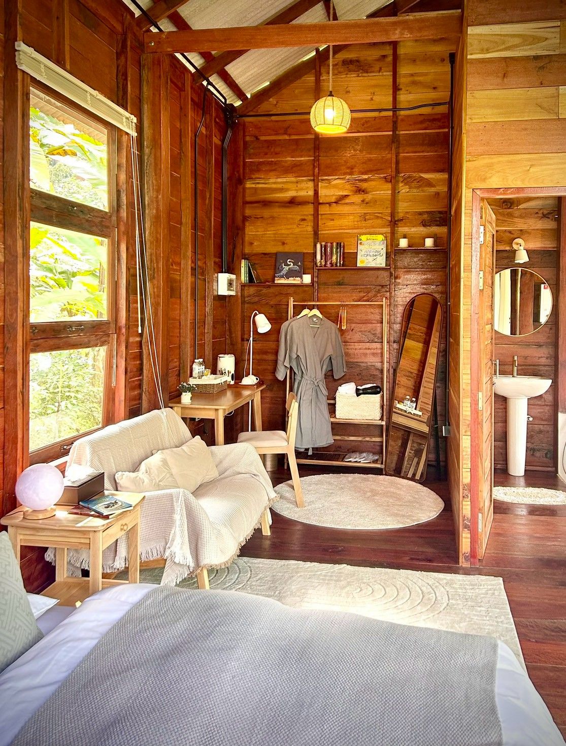 A cozy cabin interior with a bed, a desk, a chair, a hanging lamp, and a painting on the wall.