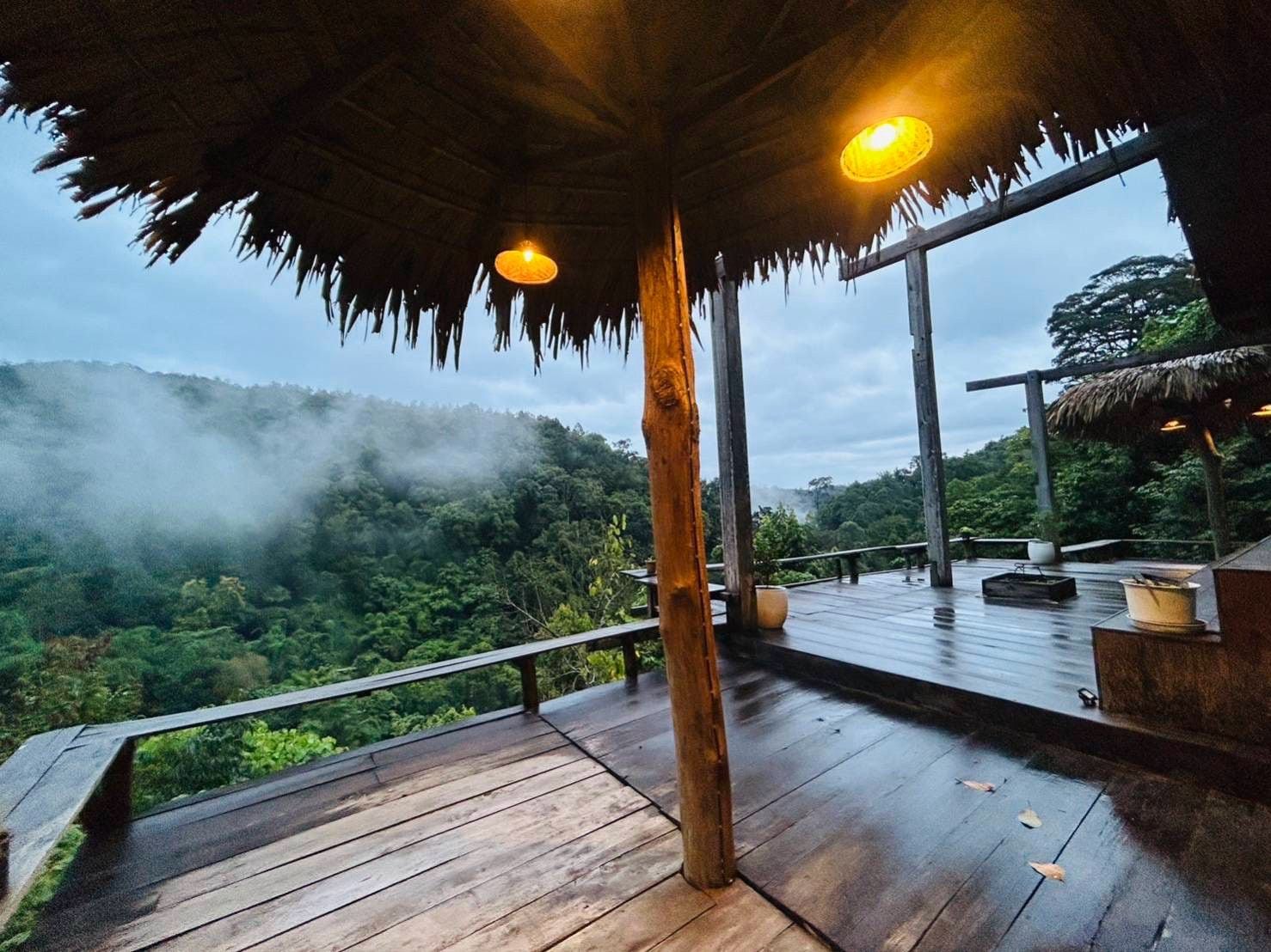 A wooden deck with a thatched roof, overlooking a lush forested landscape.