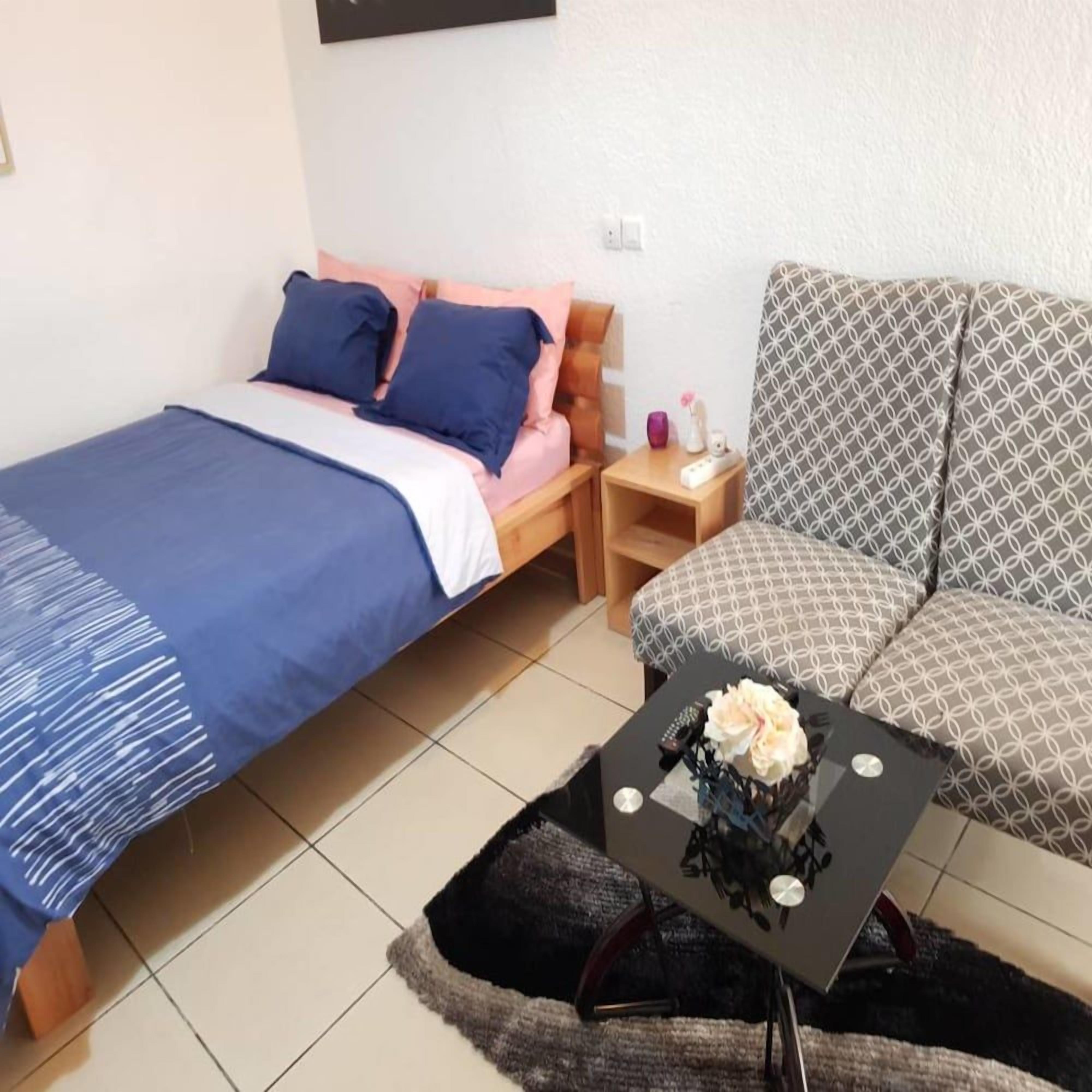 A bedroom with a bed, a bedside table, a small table with a decorative item, and a patterned sofa.