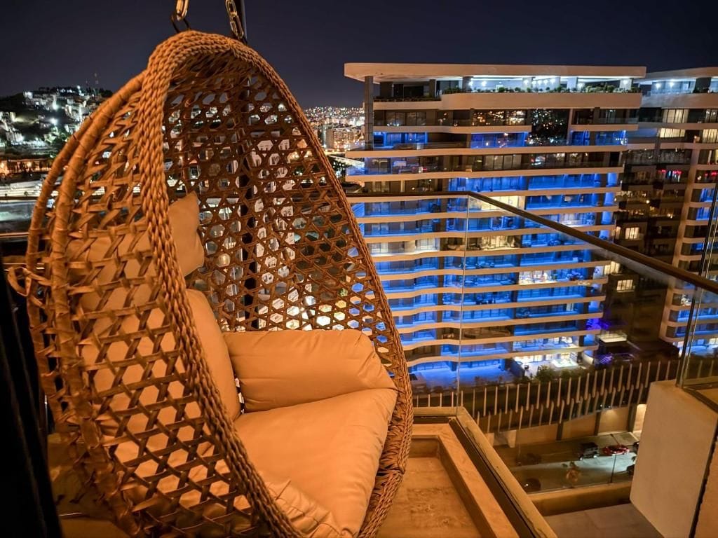 A hanging chair with a woven backrest and cushion, overlooking a cityscape with illuminated buildings.