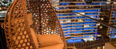 A hanging chair with a woven backrest and cushion, overlooking a cityscape with illuminated buildings.