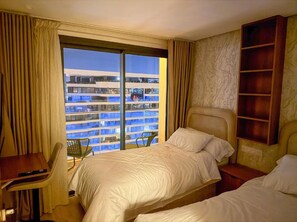 Quarto de hotel com duas camas, uma escrivaninha, uma cadeira e uma janela grande com vista para um prédio.