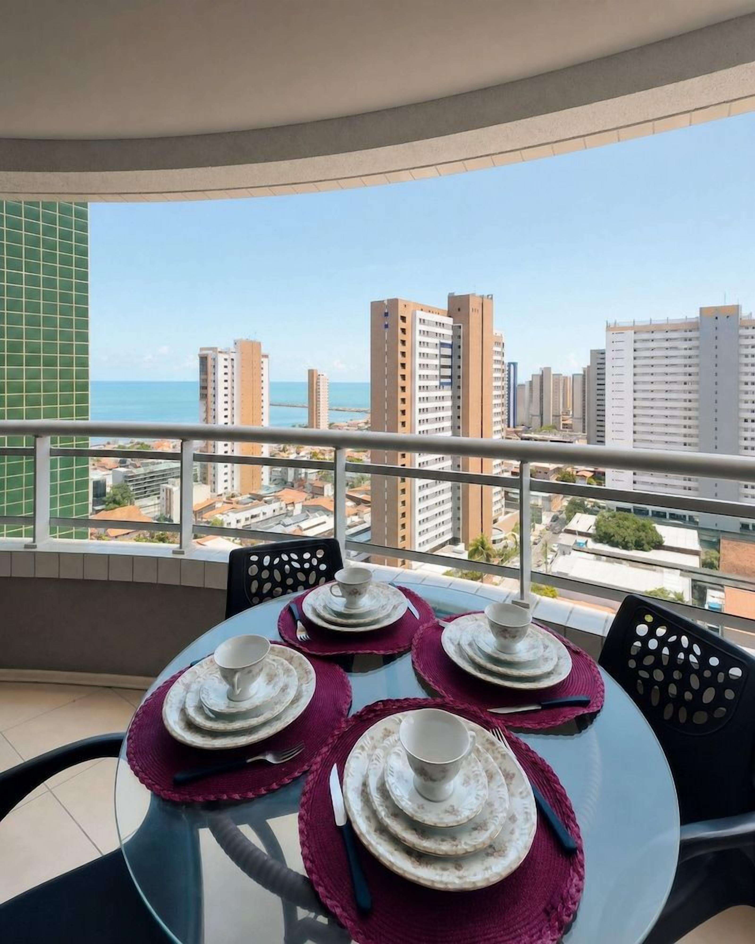 Varanda com mesa posta para refeições, vista para paisagem urbana com edifícios e um corpo d'água.