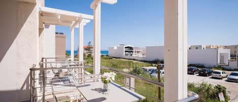 A balcony with a table and chairs, overlooking a parking area and buildings with a clear sky.