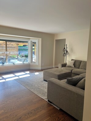 Ein geräumiges Wohnzimmer mit einem großen Fenster, einem grauen Ecksofa und einer Stehlampe.