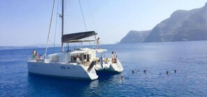 En vit yacht med ett täckt däck och en hytt, som seglar på ett lugnt hav med en bergig kustlinje i bakgrunden.