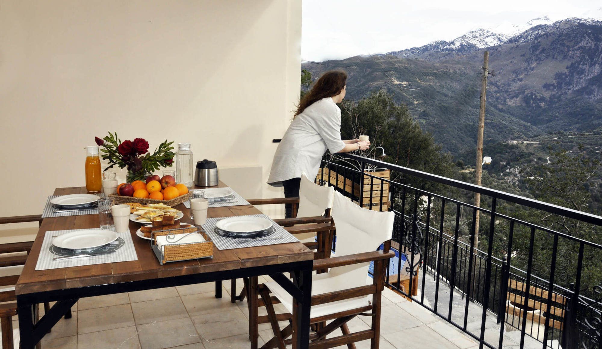 Una tavola imbandita per la colazione, una persona accanto alla ringhiera e vista su un paesaggio montuoso.