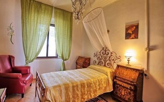 Un dormitorio con cama, una silla, una mesita de noche con lámpara y una ventana con cortinas.