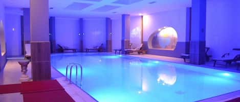 A swimming pool with a blue-lit ceiling, lounge chairs, and a decorative wall feature.