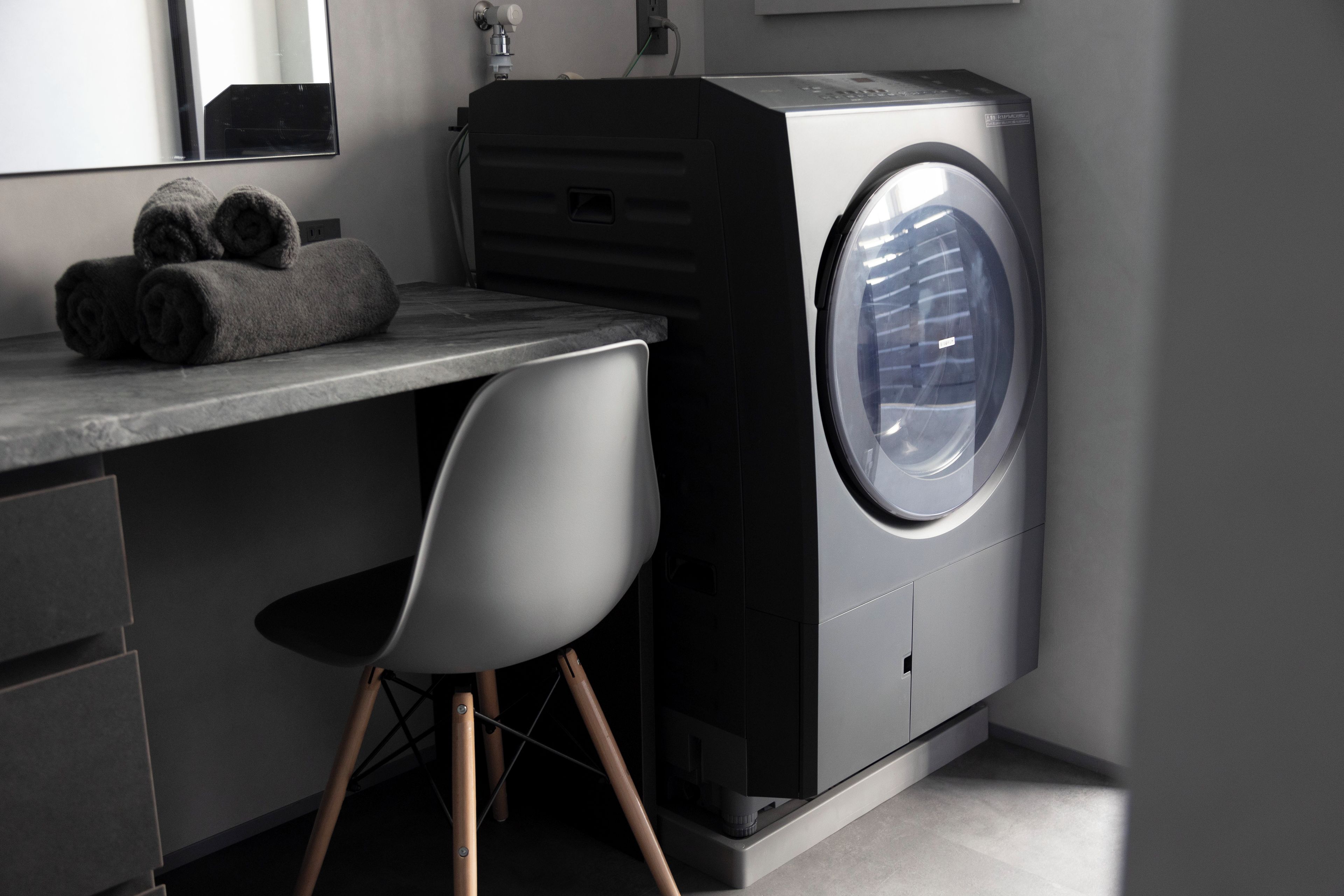A compact laundry room with a front-loading washing machine, a grey chair, and neatly folded towels on a countertop.