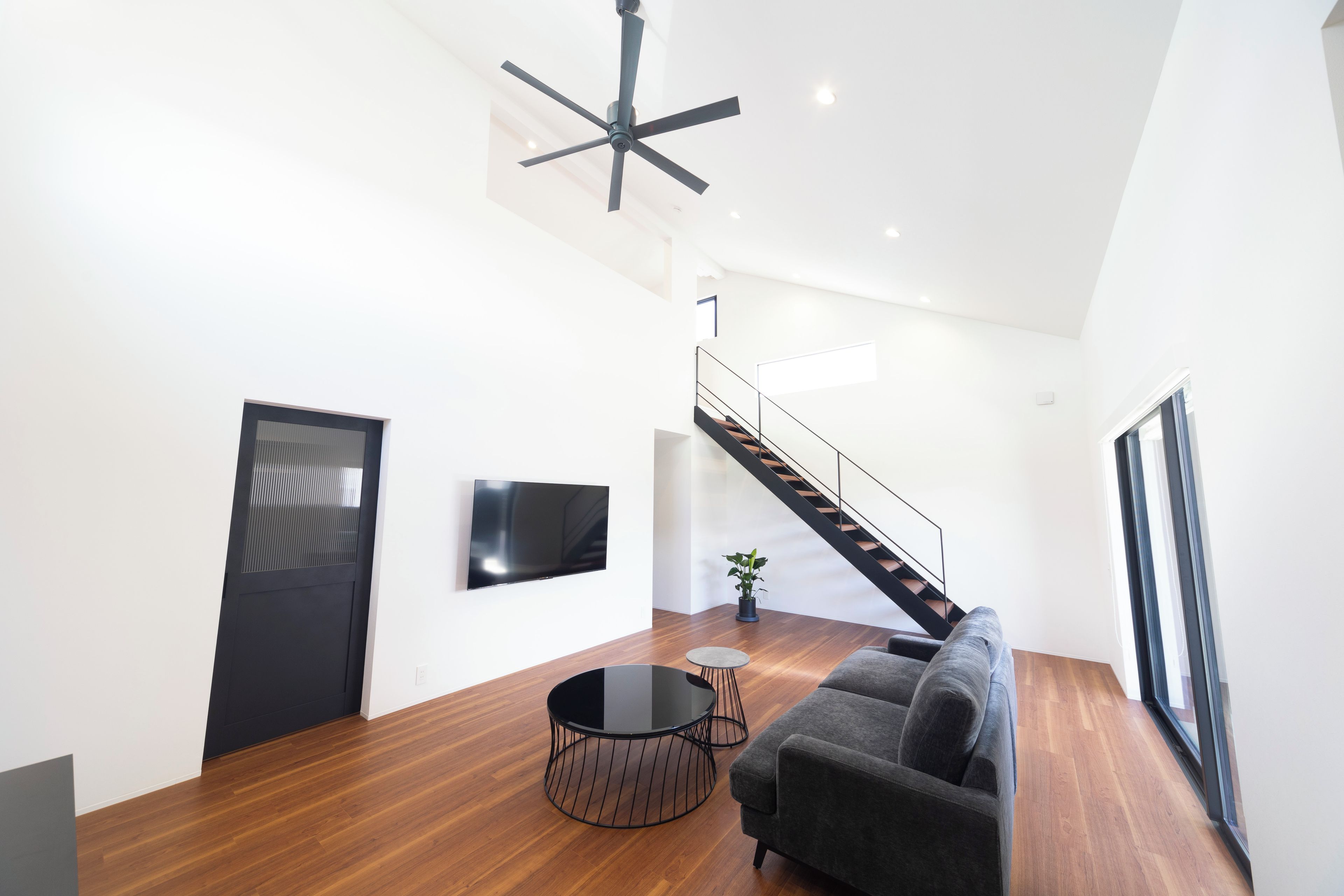 A modern living room with a flat-screen TV, a round coffee table, a grey sofa, a staircase, and a ceiling fan.