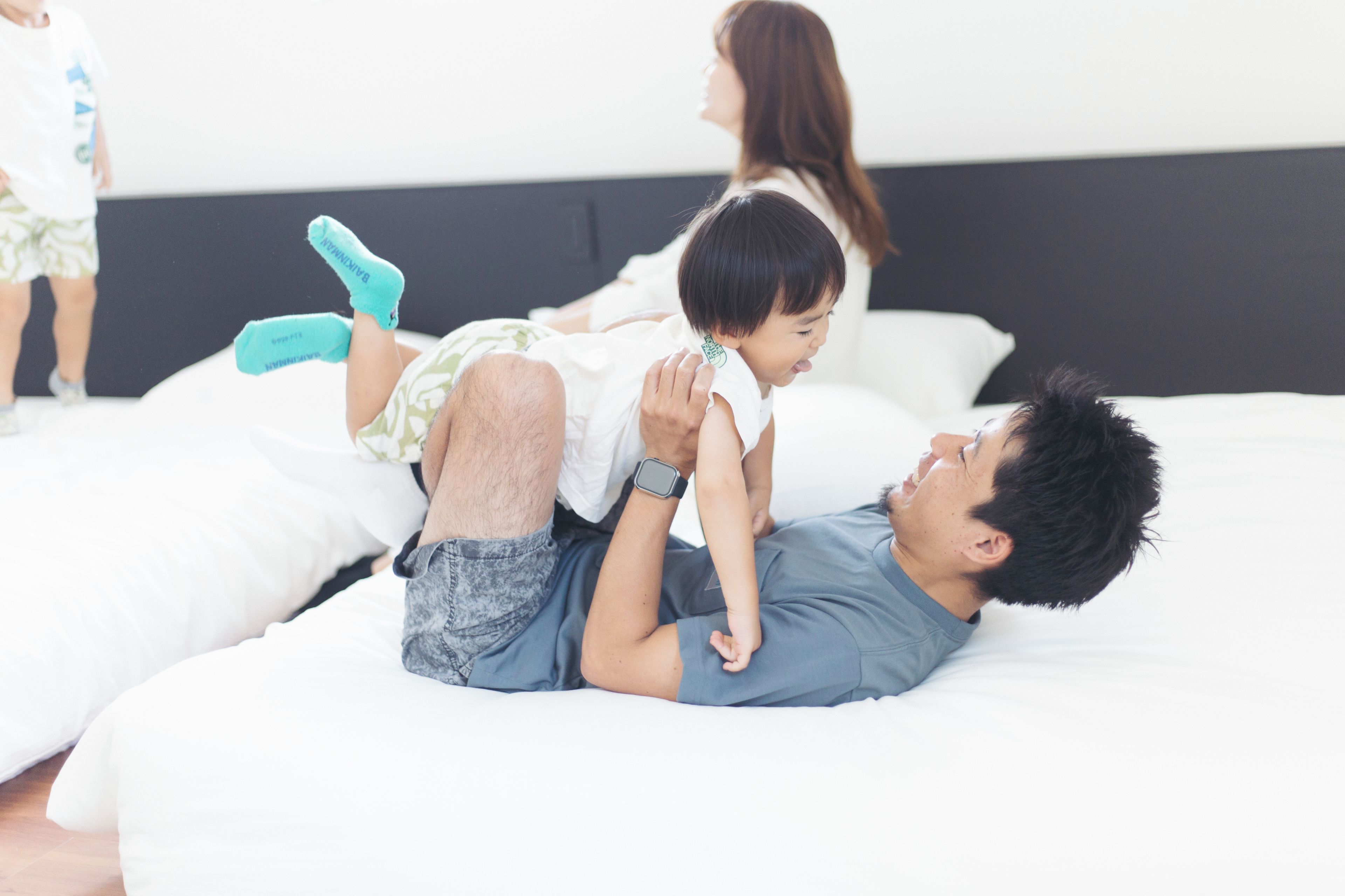 Un homme allongé sur un lit, avec un enfant sur son dos, une femme debout derrière eux et un autre enfant à l’arrière-plan.